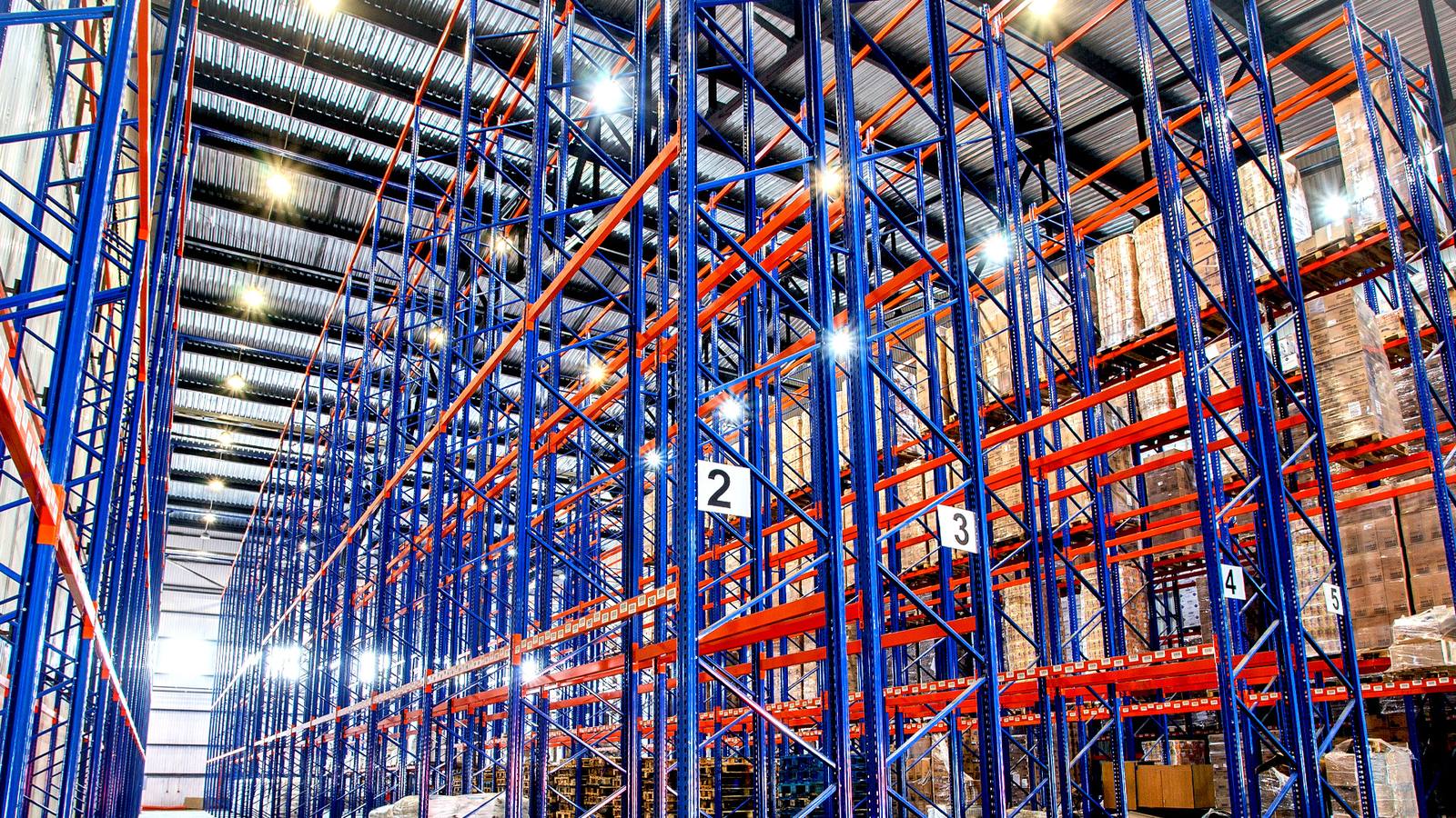 Aisles of blue and orange pallet racking, loaded and bay-numbered, in a high-bay warehouse